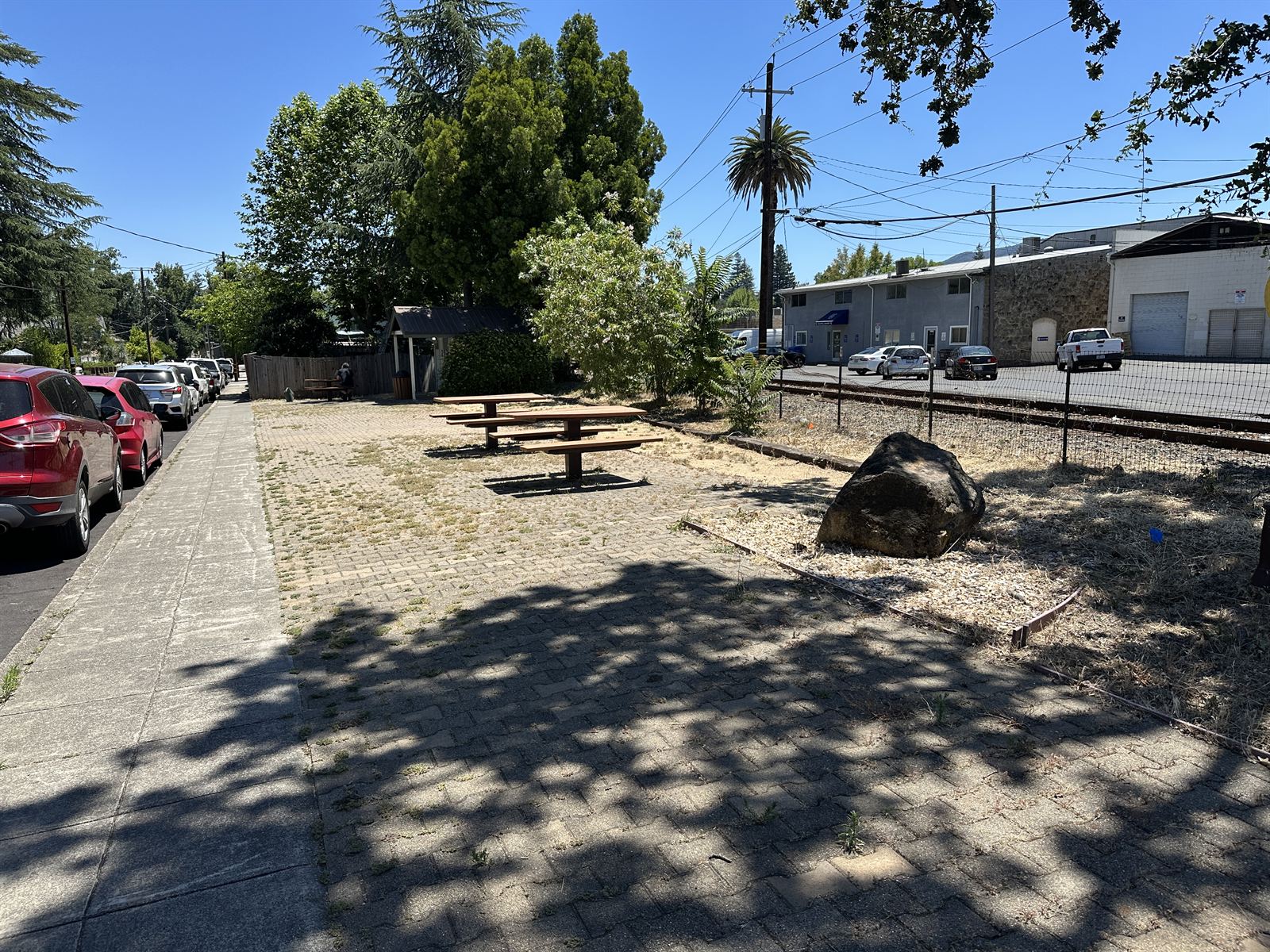 Lewis Station Park today — picnic tables, a large boulder, and mature trees on a paver courtyard beside the rail line at Church Street and Hunt Avenue in St. Helena, California
