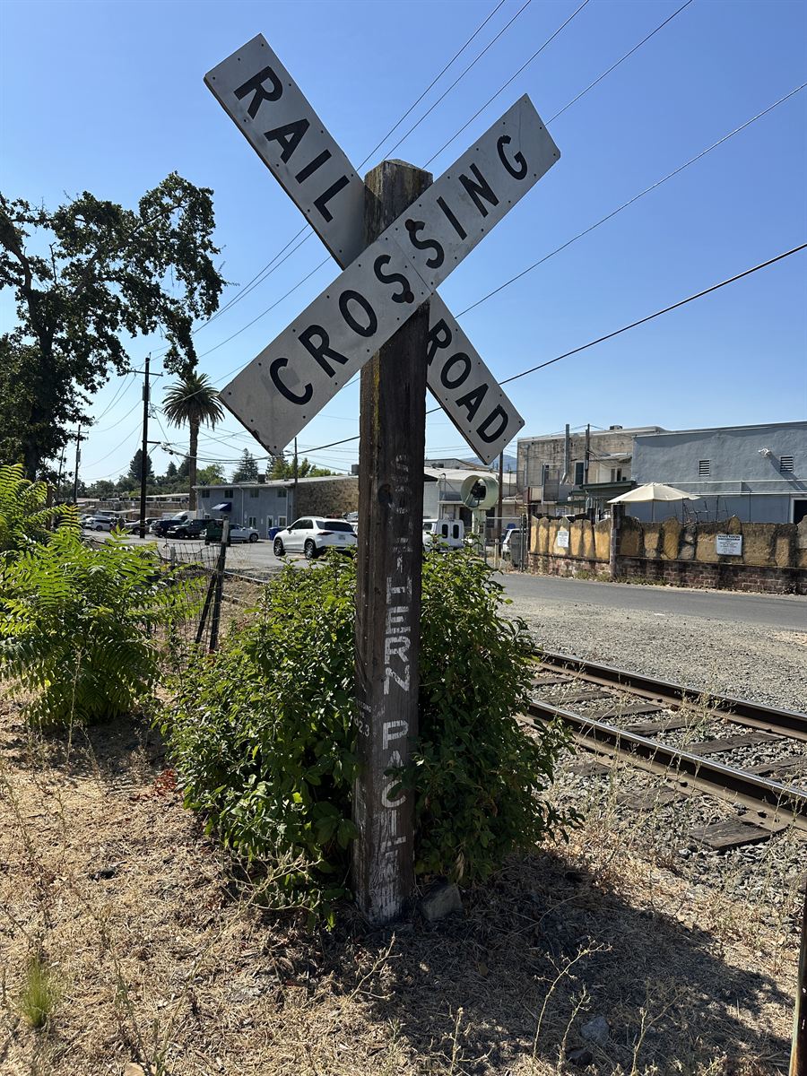 A historic Southern Pacific railroad crossing sign on a weathered wooden post at Lewis Station Park, with the rail line and downtown St. Helena behind it.