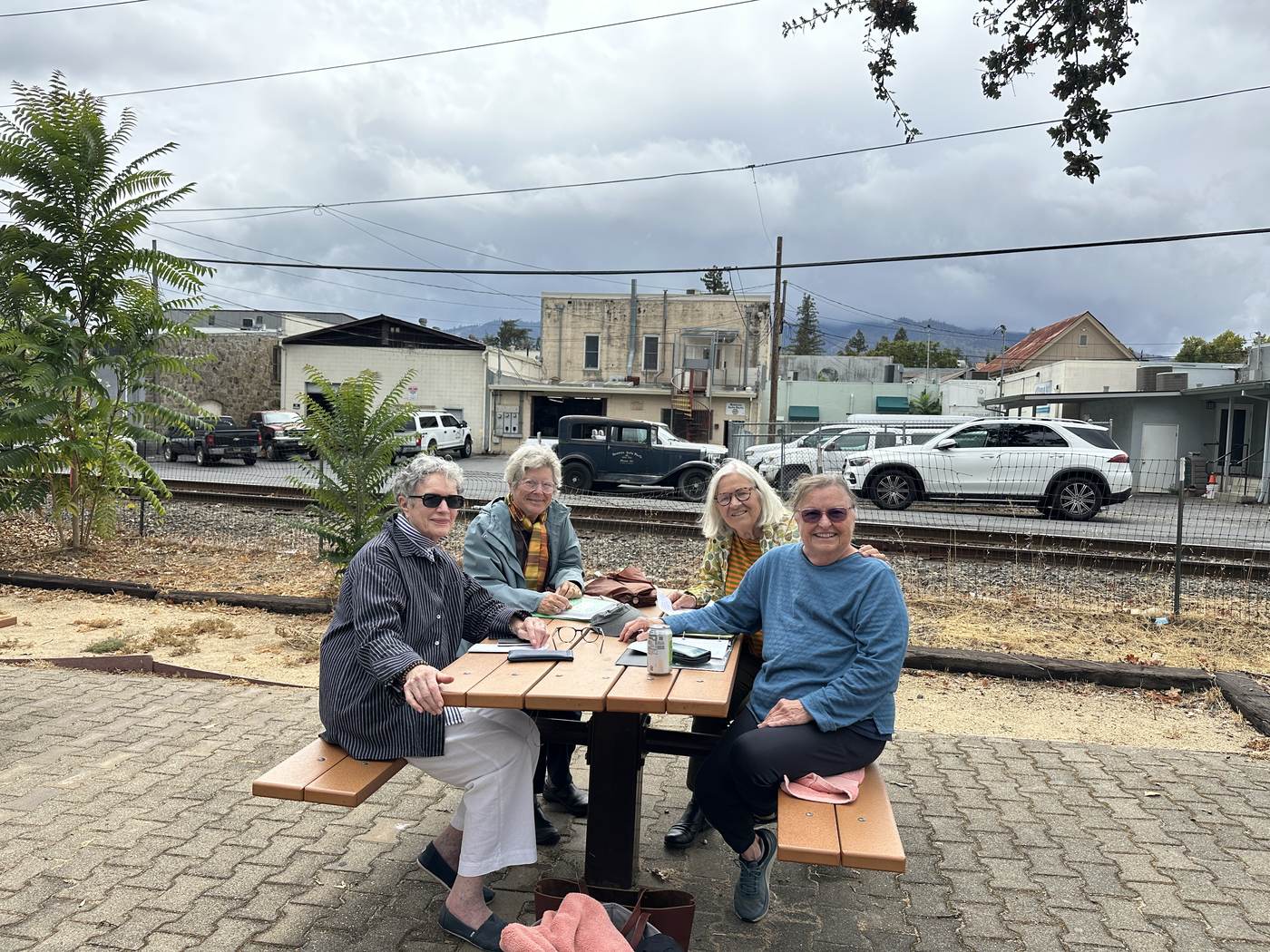 Four members of the Lewis Station Park restoration committee smiling around one of the park's picnic tables, with the rail line and downtown St. Helena behind them.