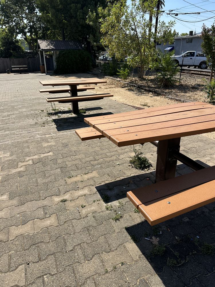 Two of the park's three picnic tables on the paver courtyard, with young trees and the rail line beyond.